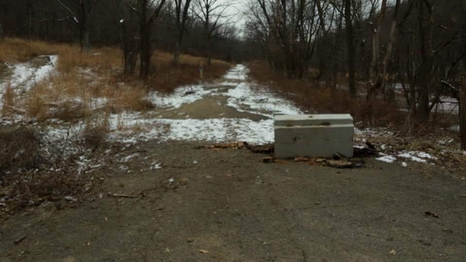 A gravel path through a wooded area, partially covered in snow, with a concrete block positioned across it to block access. Brown grass and sparse trees are visible on either side of the path, and the sky is overcast. Devou Park mountain bike trail.