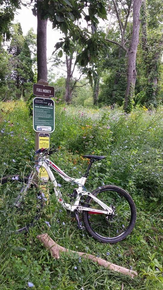 A mountain bike parked near a trail sign labeled "Full Monty," surrounded by vibrant greenery and wildflowers in a wooded area. The sign provides trail information, and a fallen log is visible in the foreground. Devou Park mountain bike trail.