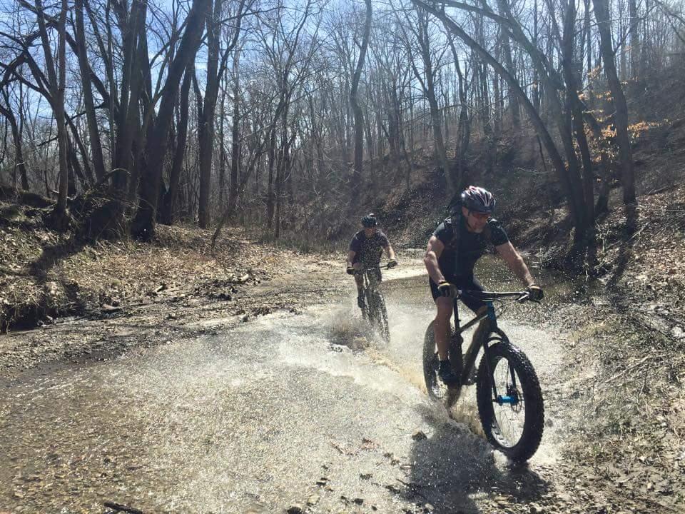 Two mountain bikers ride through a muddy trail in a forested area, splashing through water as they navigate the terrain on a sunny day. The surrounding trees are bare, indicating early spring, with a clear blue sky overhead. Caesar Creek mountain bike trail.