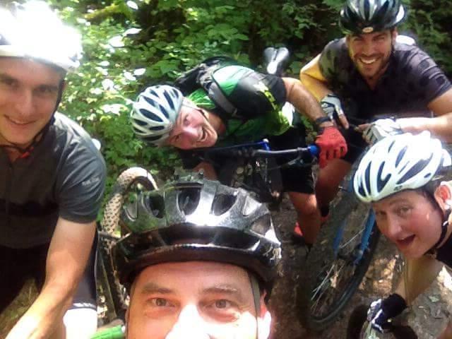 A group of five men, all wearing bicycle helmets, pose for a selfie while mountain biking in a forested area. They are surrounded by lush green trees and foliage, showcasing an outdoorsy vibe. The men are smiling and appearing to enjoy their time together. Some are holding their bikes, while others lean in closer to the camera. Caesar Creek mountain bike trail.