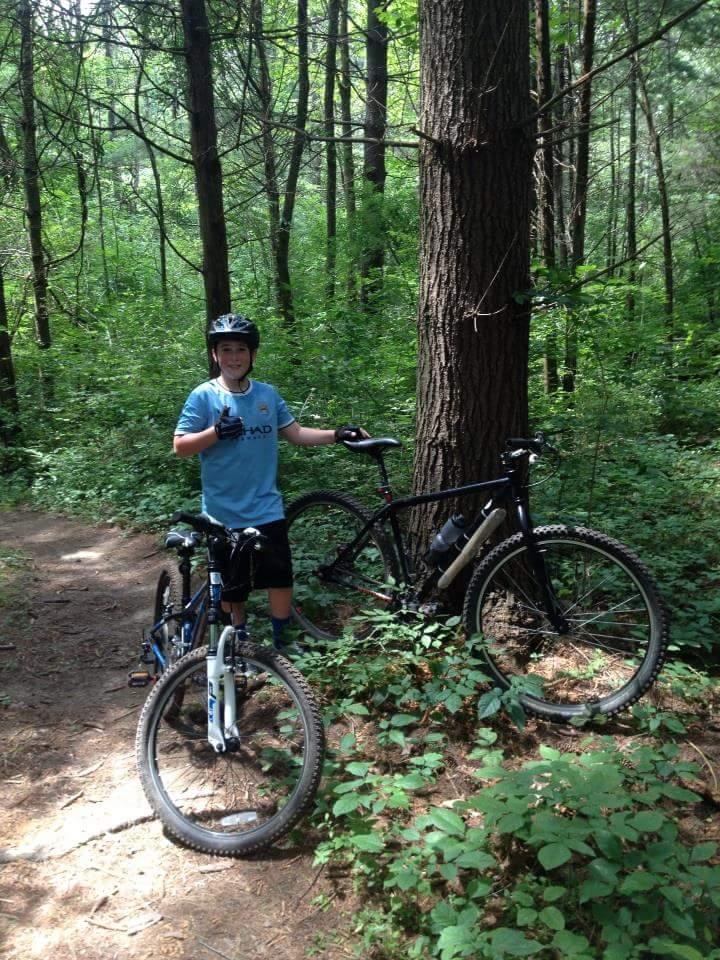 A young person wearing a blue shirt and a helmet gives a thumbs-up while standing next to two mountain bikes in a lush green forest. Trees and underbrush surround the trail where they are positioned. Caesar Creek mountain bike trail.