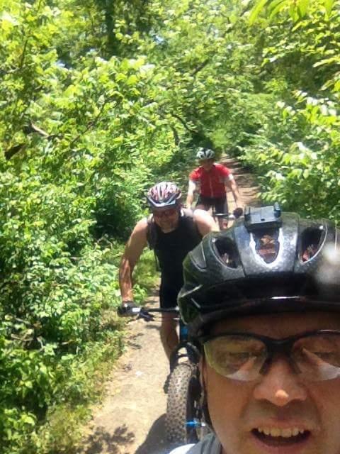 A group of three cyclists on a dirt trail surrounded by lush greenery, with the foreground showing the face and helmet of the person taking the selfie. The other two cyclists are visible in the background, enjoying their ride. Caesar Creek mountain bike trail.