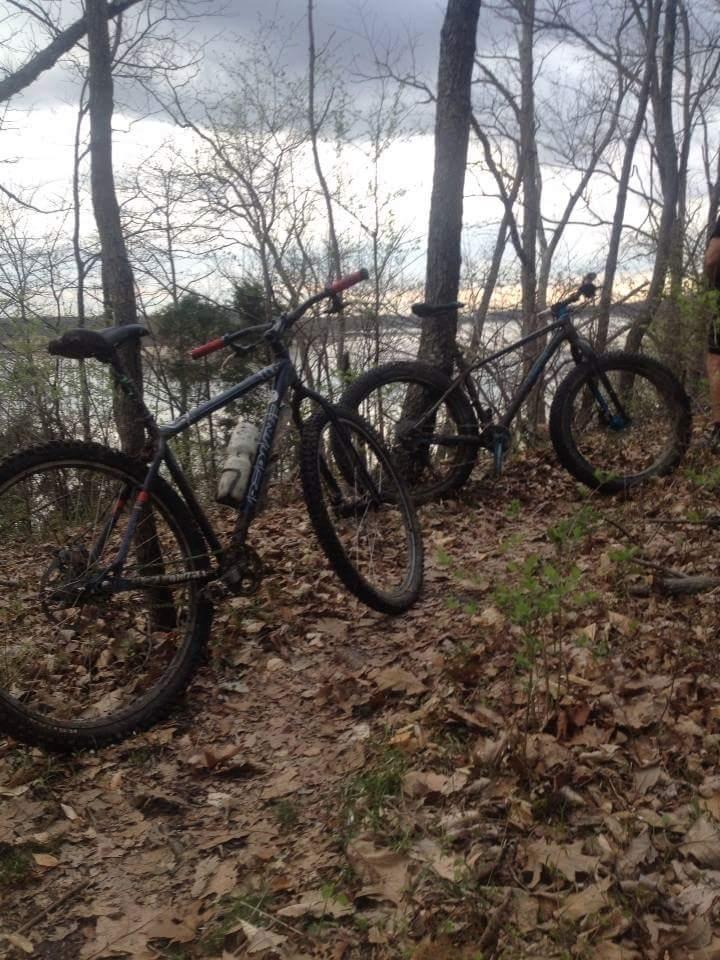 Two mountain bikes resting on a leaf-covered trail in a wooded area, with trees and a body of water visible in the background under a cloudy sky. Caesar Creek mountain bike trail.