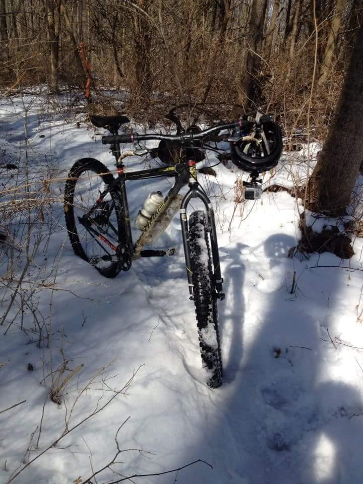 A mountain bike resting on a snow-covered trail surrounded by bare trees and brush. The bike has snow on its tires and a water bottle attached to the frame, indicating it's been used in a winter outdoor setting. Sunlight filters through the trees, casting shadows on the ground. MoMBA @ Huffman MetroPark mountain bike trail.
