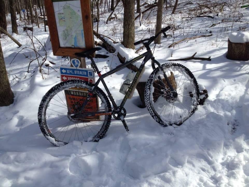 A mountain bike with snow-covered tires is parked beside a trail map and signage in a snowy forest setting. Trees are visible in the background, and the ground is blanketed in fresh snow. MoMBA @ Huffman MetroPark mountain bike trail.