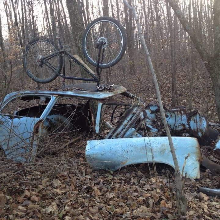 An abandoned, rusty blue car partially covered in foliage, with a black bicycle upside down resting on top of it, set in a wooded area during the fall season. Caesar Creek mountain bike trail.