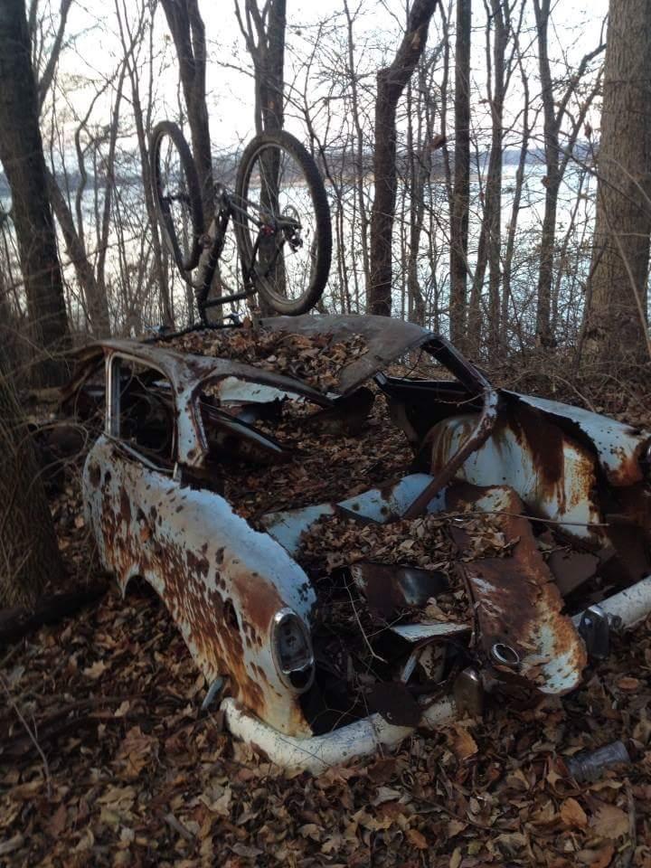 An abandoned, rusted car covered in leaves, with a bicycle resting upside down on top, surrounded by trees and a glimpse of water in the background. Caesar Creek mountain bike trail.
