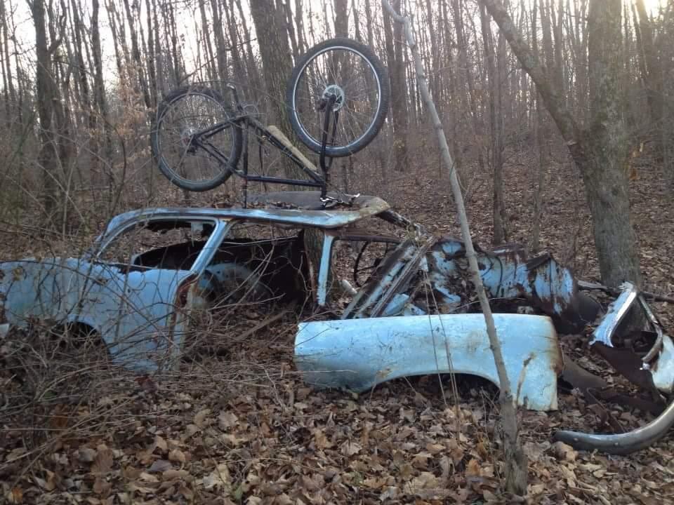A rusted blue car partially submerged in foliage and surrounded by trees, with a bicycle resting upside down on its roof. The scene captures a blend of nature and abandoned objects in a wooded area during daytime. Caesar Creek mountain bike trail.