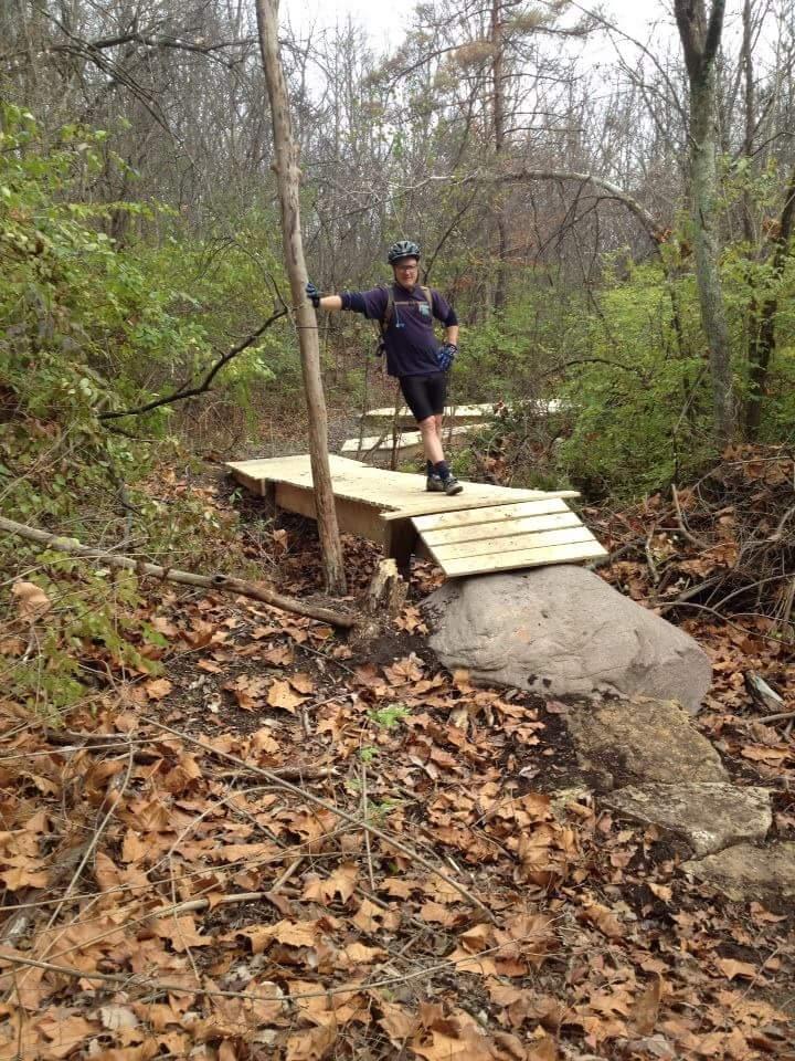 A person in a helmet and cycling gear stands on a wooden bridge that spans a rocky area in a wooded setting. Surrounding the bridge are leaf-covered ground and various trees, indicating a natural outdoor environment. The individual leans against a tree while posing confidently. MoMBA @ Huffman MetroPark mountain bike trail.