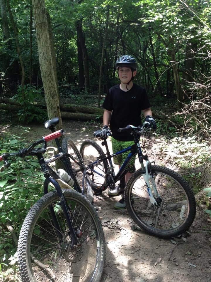 A young person wearing a black t-shirt and a helmet stands beside two mountain bikes in a wooded area. The scene features lush green trees and undergrowth, suggesting a nature trail. MoMBA @ Huffman MetroPark mountain bike trail.