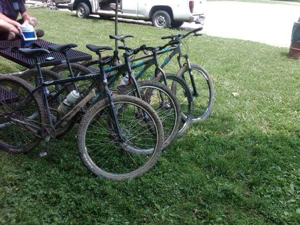 Four mountain bikes are lined up on a grassy area, showing signs of use with some dirt on the tires. In the background, a parked white pickup truck is visible, along with a picnic table where a person is seated, holding a drink. MoMBA @ Huffman MetroPark mountain bike trail.