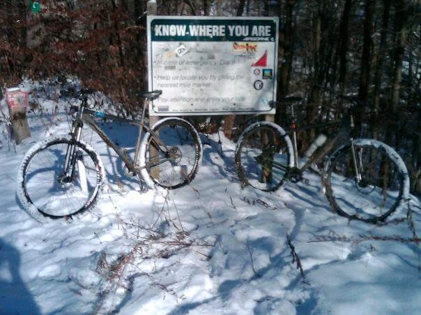 Two mountain bikes are parked in a snowy landscape in front of a trail sign. The sign features important information about navigation and emergency contact. Snow covers the ground and clings to the bikes’ tires. Trees are visible in the background, suggesting a forested area. Caesar Creek mountain bike trail.