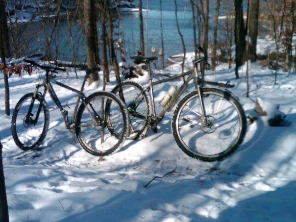 Two mountain bikes parked in a snowy forest setting, with a glimpse of a blue lake visible in the background. The bikes are surrounded by snow-covered ground and tall trees. Caesar Creek mountain bike trail.