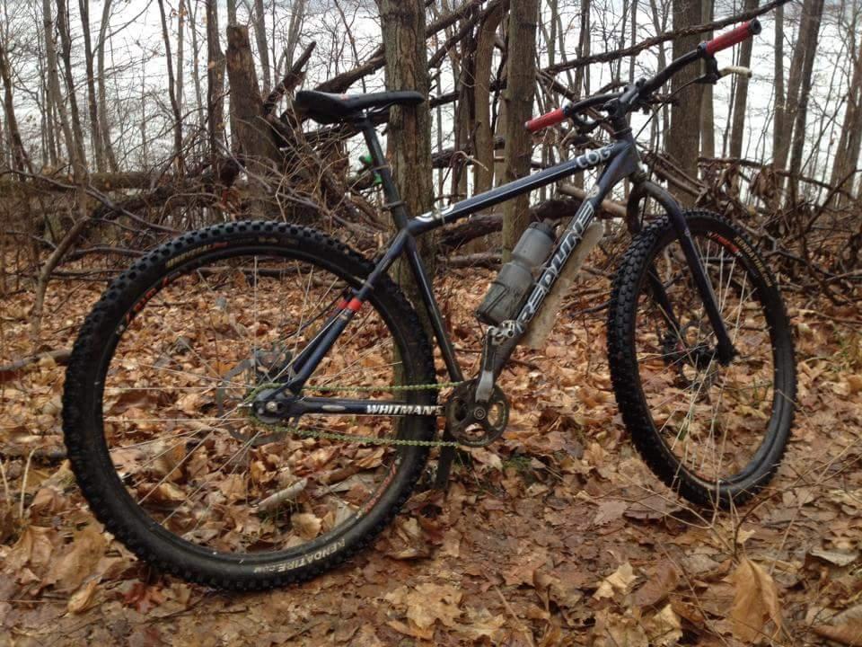 A mountain bike positioned on a forest floor covered with fallen leaves. The bike features thick, knobby tires suitable for off-road terrain, a sturdy frame, and a water bottle mounted on the side. In the background, there are bare trees and branches, indicating a wooded area in early spring or late fall. Caesar Creek mountain bike trail.