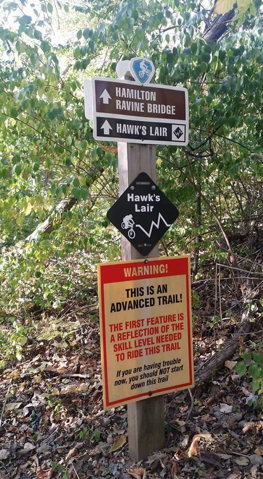 A wooden signpost in a wooded area featuring directional signs for "Hamilton Ravine Bridge" and "Hawk's Lair." Below, a warning sign indicates "THIS IS AN ADVANCED TRAIL!" with additional information about skill level requirements for riding the trail. The signs are surrounded by trees and foliage. MoMBA @ Huffman MetroPark mountain bike trail.