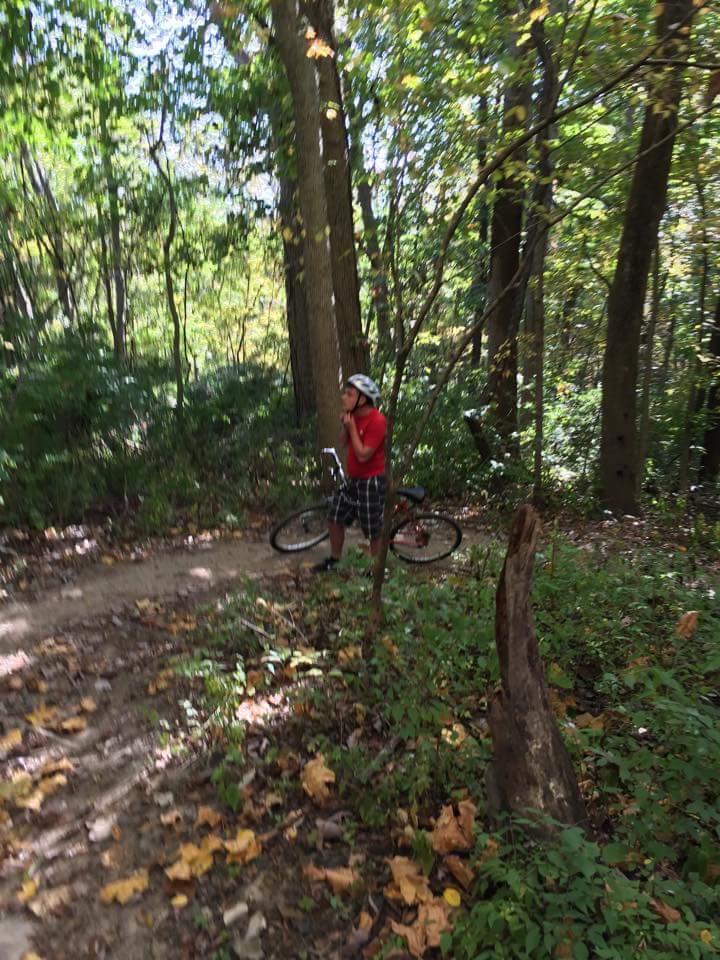 A person in a red shirt and black and white plaid shorts stands beside a mountain bike on a dirt path surrounded by trees. The scene captures a wooded area with fallen leaves on the ground, indicating autumn. MoMBA @ Huffman MetroPark mountain bike trail.