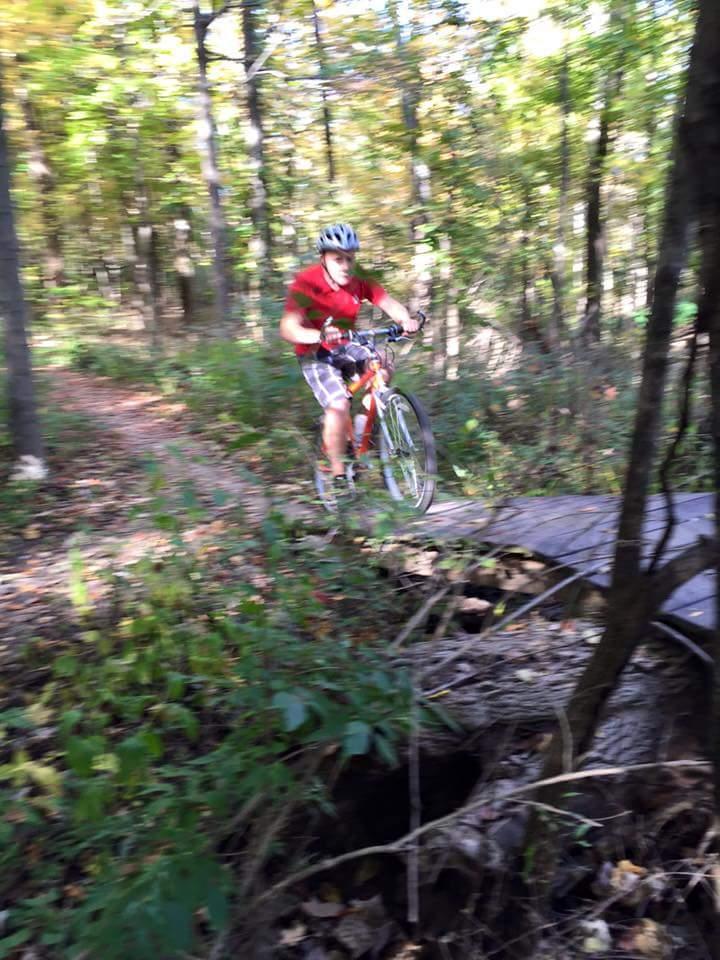 A cyclist in a red shirt and helmet rides over a small wooden bridge on a forest trail, surrounded by colorful autumn foliage. The image is slightly blurred, capturing the motion and speed of the ride. MoMBA @ Huffman MetroPark mountain bike trail.