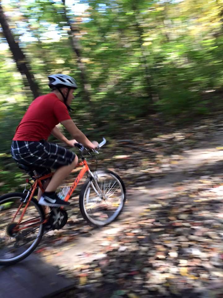 A person wearing a helmet and a red shirt rides a mountain bike along a wooded trail, surrounded by colorful autumn leaves. The image has a motion blur effect, conveying a sense of speed and movement. MoMBA @ Huffman MetroPark mountain bike trail.