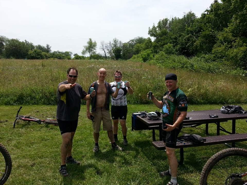 Four men standing together in a grassy outdoor area, celebrating after a biking session. They are smiling and holding drinks, with mountain bikes parked nearby. A picnic table is visible in the background, surrounded by trees and tall grass. MoMBA @ Huffman MetroPark mountain bike trail.