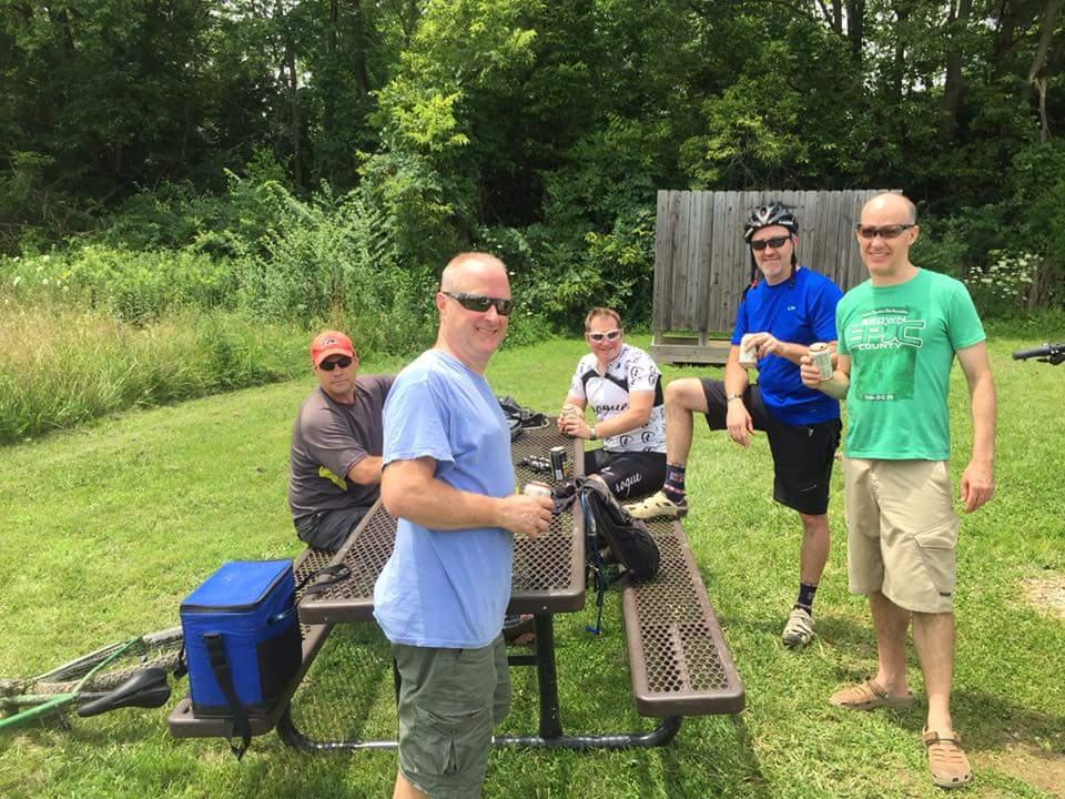 Group of five men enjoying a break outdoors during a biking trip, sitting around a picnic table in a lush green area. They are casually dressed, with some wearing sunglasses and one man in cycling gear. Drinks are in their hands, and bicycles are leaned against the table. MoMBA @ Huffman MetroPark mountain bike trail.