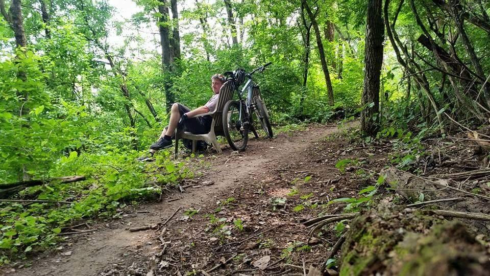 A person sitting on a bench along a dirt trail in a lush green forest, with a bicycle resting beside them. The scene captures the tranquility of nature with dense foliage surrounding the area. MoMBA @ Huffman MetroPark mountain bike trail.