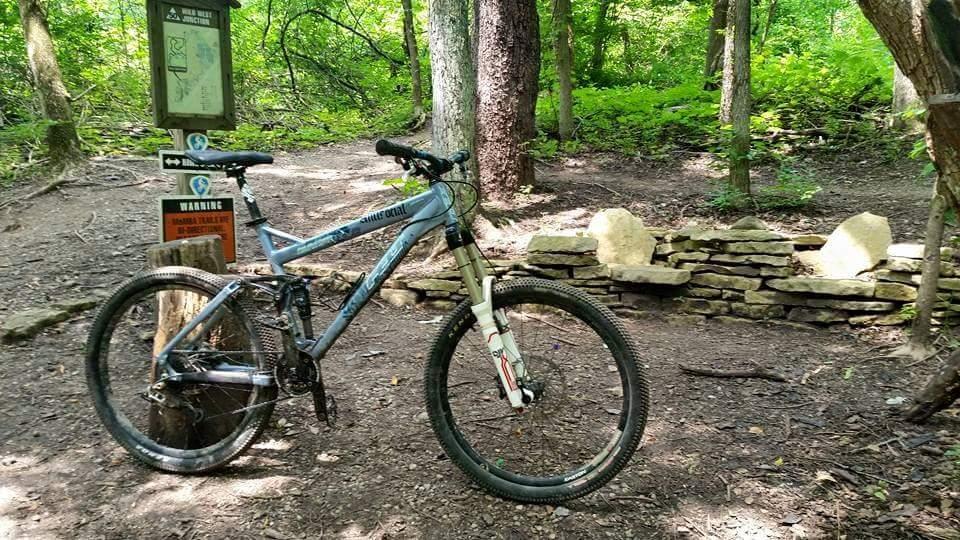 A mountain bike resting on a dirt path in a lush green forest. In the background, there are trail signs and a stone seating area. The scene conveys a tranquil outdoor setting ideal for biking and hiking activities. MoMBA @ Huffman MetroPark mountain bike trail.