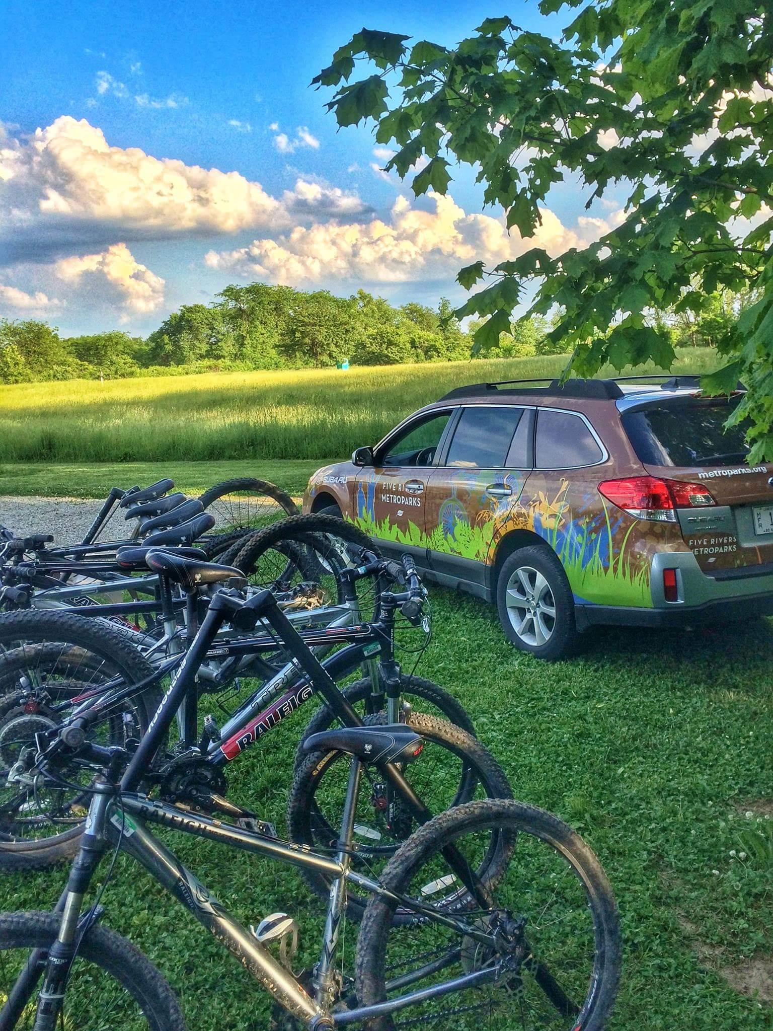 A parked vehicle with a nature-themed design is beside a cluster of mountain bikes on grass. In the background, a lush, green landscape stretches under a bright blue sky, dotted with fluffy white clouds. MoMBA @ Huffman MetroPark mountain bike trail.