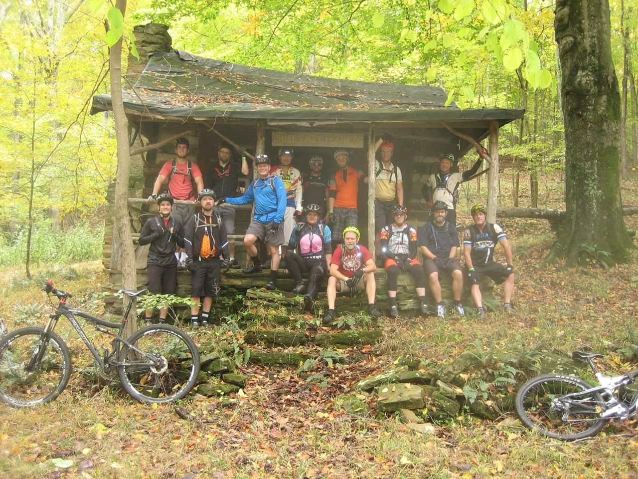 A group of 15 mountain bikers gathered on the steps in front of a rustic wooden shelter surrounded by autumn foliage. Some of the bikers are wearing helmets and cycling gear, while mountain bikes are parked alongside. The scene captures a vibrant outdoor atmosphere. Brown County Park mountain bike trail.