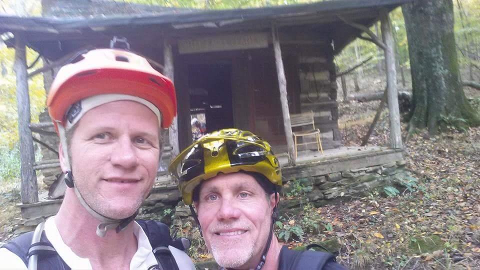 Two smiling individuals in mountain biking gear pose for a selfie in front of a rustic log cabin surrounded by trees and autumn foliage. The cabin has a wooden porch, and a chair is visible on it, while the ground is covered in leaves. Brown County Park mountain bike trail.