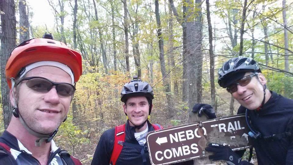 Three mountain bikers smiling for a selfie in a forest, standing in front of a wooden trail sign that points left towards "Nebo Trail" and "Combs." The trees in the background show autumn foliage. Each cyclist is wearing a helmet and sunglasses. Brown County Park mountain bike trail.