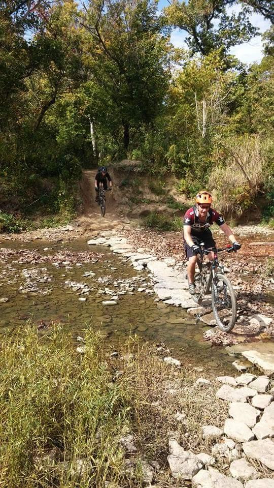 Two mountain bikers ride along a rocky trail that crosses a shallow creek. One biker is navigating stones that create a path over the water, while another biker approaches from a dirt path in the background. The scene is surrounded by lush green trees under a clear blue sky, showcasing a vibrant autumn landscape. Versailles State Park mountain bike trail.