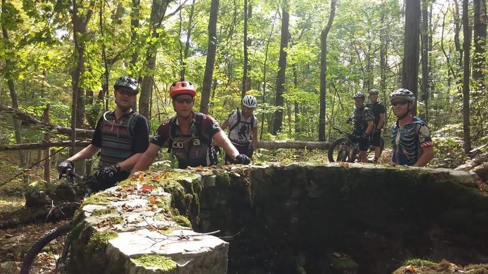 A group of six mountain bikers gathered around a moss-covered stone structure in a wooded area. The scene features vibrant green trees in the background, with sunlight filtering through the leaves. The riders are wearing helmets and cycling gear, appearing to engage with one another as they explore the trail. Versailles State Park mountain bike trail.