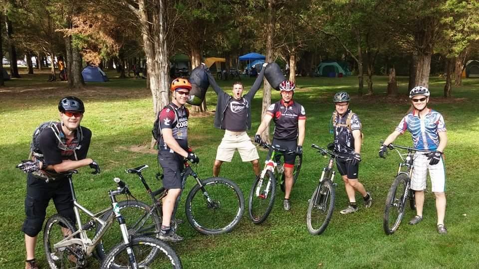 A group of six mountain bikers poses together in a grassy area surrounded by trees. They are smiling and wearing biking gear, including helmets and jerseys. Two cyclists are holding their bikes, while one is playfully jumping in the background. In the distance, tents can be seen, indicating a camping setup. The scene conveys a sense of camaraderie and outdoor adventure. Versailles State Park mountain bike trail.