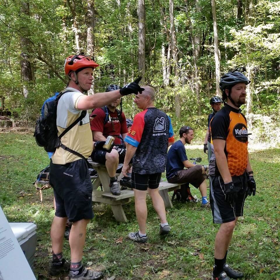 A group of mountain bikers gathered in a wooded area, discussing a trail. One cyclist in an orange helmet is pointing and talking, while others are seated on a picnic table or standing nearby, wearing various biking jerseys. The scene is surrounded by lush greenery, indicating a day of outdoor activity. Versailles State Park mountain bike trail.
