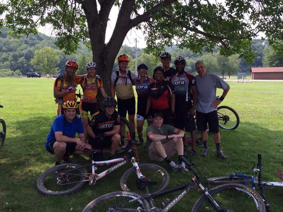 A group of cyclists gathered under a tree in a park, smiling and posing for a photo. Some are wearing colorful biking jerseys and helmets, while their mountain bikes are resting on the grass nearby. The background features green hills and a few buildings. The scene captures a fun, outdoor biking community event. Versailles State Park mountain bike trail.