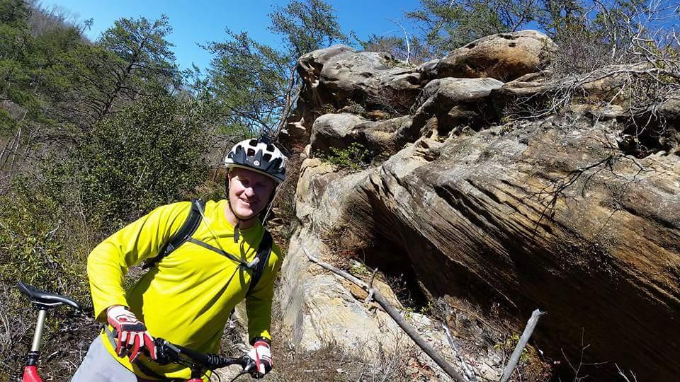 A person wearing a yellow long-sleeve shirt and a bicycle helmet smiles while standing next to a large rock formation in a wooded area. Sunlight shines through trees in the background, indicating a clear day. A mountain bike is leaning beside them. Big South Fork mountain bike trail.