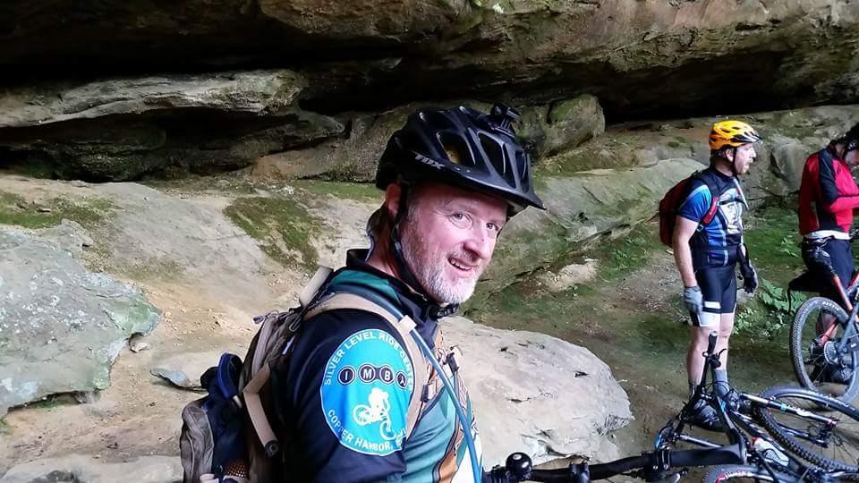 A group of mountain bikers resting in a natural cave-like area. One man, wearing a blue and green cycling jersey with a helmet, is smiling at the camera, while two other cyclists in the background appear to be adjusting their gear. The rocky surroundings are partially covered in greenery, indicating an outdoor setting. Big South Fork mountain bike trail.