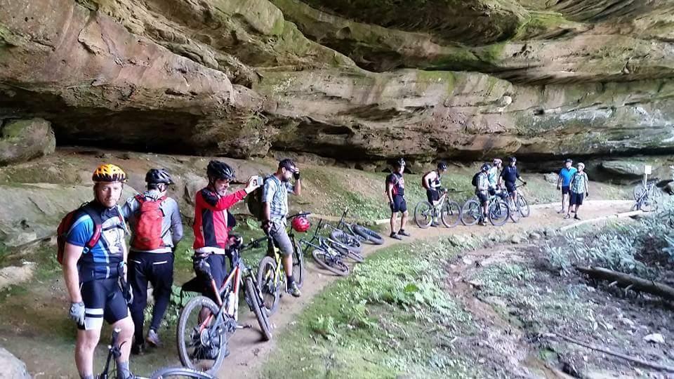 A group of mountain bikers pauses along a dirt trail near a rocky overhang, with their bikes resting beside them. The scene features lush greenery and a natural cave-like formation in the background. Some riders are adjusting their gear while others are taking photos or chatting. The group showcases a mix of cycling attire and helmets, emphasizing an outdoor recreational activity in a scenic, forested area. Big South Fork mountain bike trail.