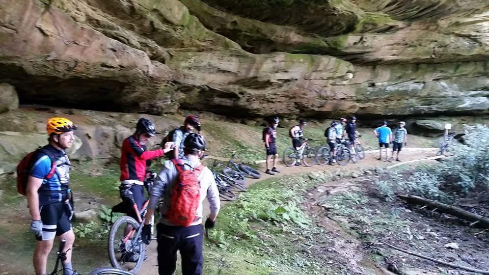 A group of mountain bikers gathered at the entrance of a rocky cave, surrounded by lush greenery. Some are adjusting their gear while others converse. Mountain bikes are parked along the winding trail, which leads deeper into the forested area. The scene captures the camaraderie and adventure spirit of outdoor biking in a natural landscape. Big South Fork mountain bike trail.