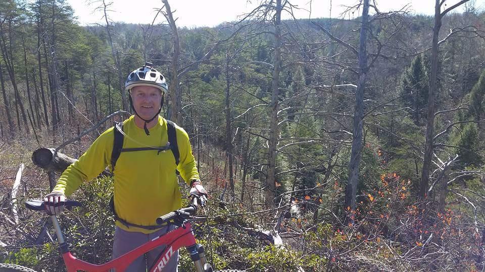 A smiling person wearing a yellow long-sleeve shirt and a helmet stands next to a red mountain bike on a wooded hillside. Behind them are tall, bare trees and a lush green landscape in the distance, indicating a sunny day ideal for outdoor activities. Big South Fork mountain bike trail.
