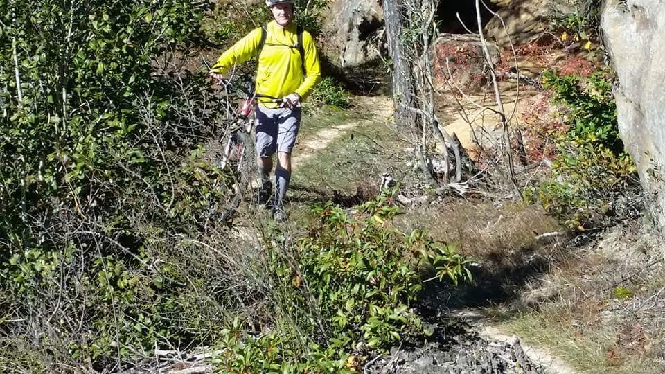A cyclist navigating a narrow, overgrown trail in a natural setting, wearing a bright yellow long-sleeve shirt, gray shorts, and a helmet. The path is surrounded by dense vegetation and rocky formations. Big South Fork mountain bike trail.