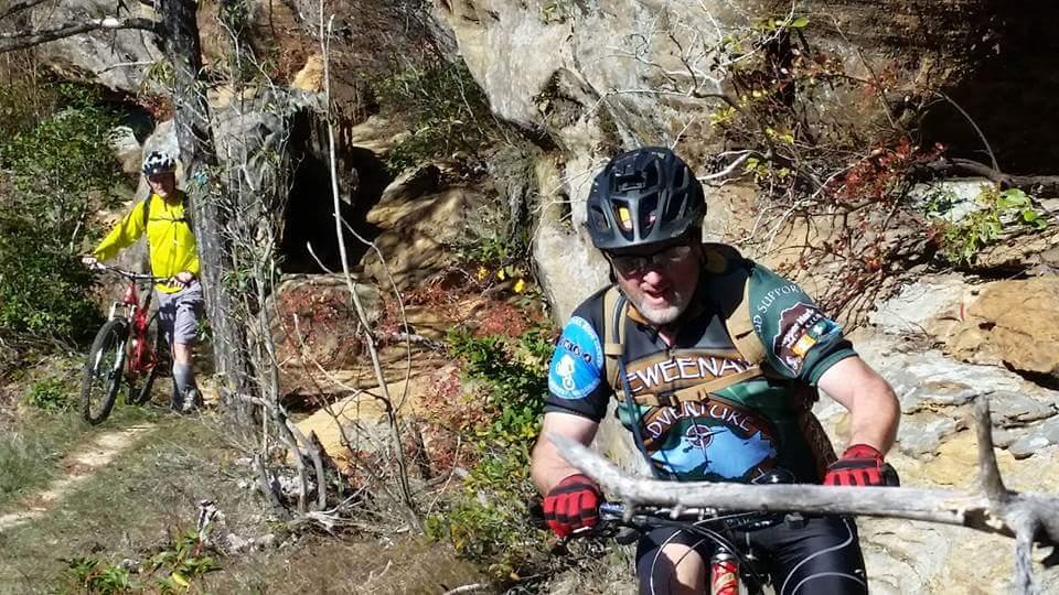 Two mountain bikers navigate a rugged trail, with one rider in a colorful jersey pulling a stick from the path while the other, wearing a yellow jacket, is walking beside their bike. The scene is set against a backdrop of rocky terrain and lush vegetation, showcasing a sunny day on a nature trail. Big South Fork mountain bike trail.