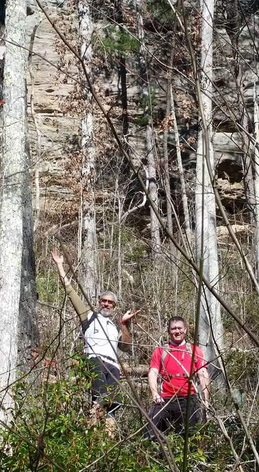 Two hikers pose for a photo in a forested area, surrounded by tall trees and underbrush. One hiker is wearing a white shirt and has a playful gesture with one arm raised, while the other is in a red shirt and smiling. The background features a rocky hillside. Big South Fork mountain bike trail.