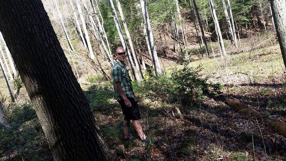 A person standing in a forested area, surrounded by tall trees and underbrush. The individual is wearing a short-sleeved checked shirt and shorts, and is looking back toward the camera while standing on a leaf-covered ground. Sunlight filters through the trees, creating a natural and serene atmosphere. Big South Fork mountain bike trail.