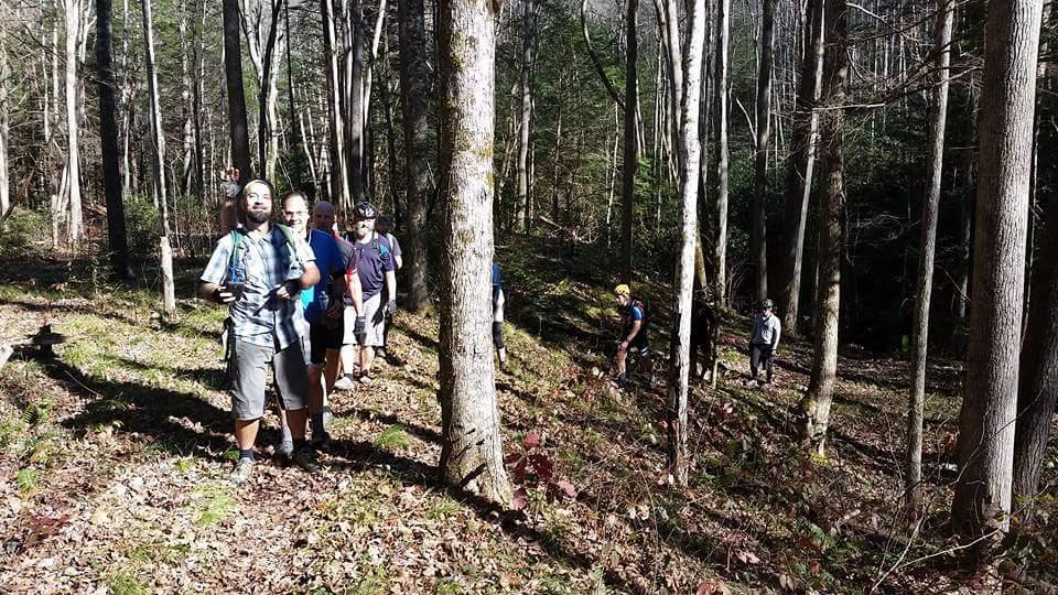 A group of hikers walking on a forest trail surrounded by tall trees and greenery. The sunlight filters through the leaves, creating a natural setting. Some hikers are smiling and interacting, while others are focused on the trail ahead. Big South Fork mountain bike trail.