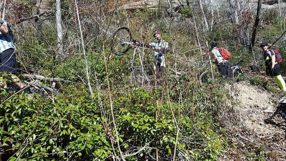 A group of mountain bikers navigating a rugged, overgrown trail surrounded by trees and shrubs. Some riders are seen lifting their bikes while others stand nearby, assessing the path ahead. The scene captures the challenges of off-road biking in a natural environment. Big South Fork mountain bike trail.
