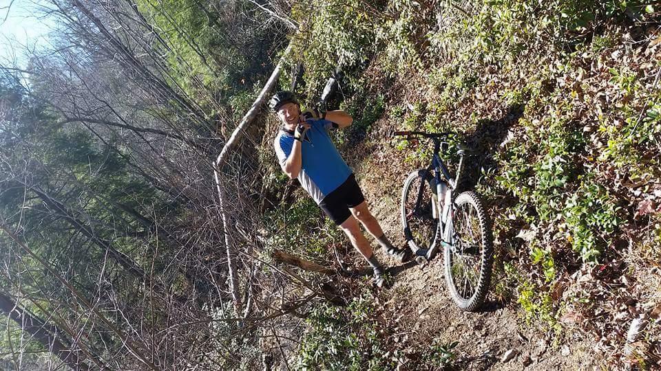 A person wearing a helmet and athletic clothing stands next to a mountain bike on a forest trail, surrounded by trees and foliage. The individual is smiling and appears to be enjoying the outdoor setting. Big South Fork mountain bike trail.