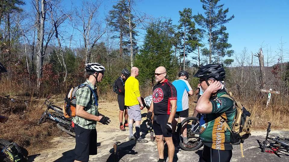 A group of mountain bikers in outdoor gear gather on a rocky trail surrounded by trees. Some cyclists are adjusting their helmets and chatting, while others are preparing their bikes. The scene is set on a sunny day with a clear blue sky. Big South Fork mountain bike trail.