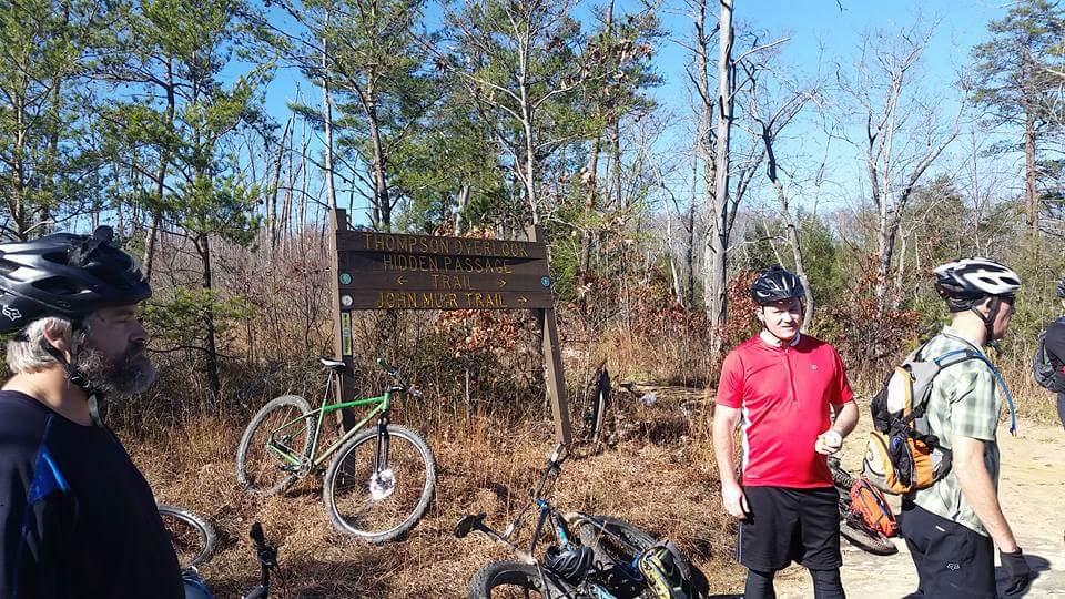 A group of three mountain bikers stands near a trail sign indicating the Thompson DeRosa Hidden Passage Trail and John Muir Trail. Two bicycles are parked in the background, surrounded by a natural setting of trees and dry grass. The scene is bright and sunny, with clear blue skies. Big South Fork mountain bike trail.