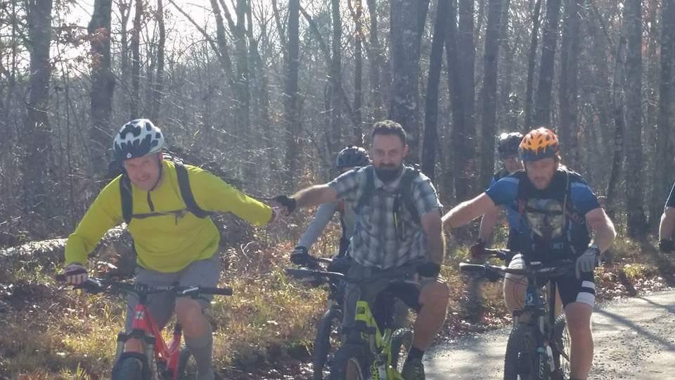 Four mountain bikers are riding on a trail through a wooded area. One cyclist in a bright yellow shirt is in the foreground, smiling and pedaling hard, while another with a beard is next to him, extending his hand. The other two bikers follow behind, all wearing helmets and engaged in the activity. Sunlight filters through the trees, casting a warm glow on the scene. Big South Fork mountain bike trail.